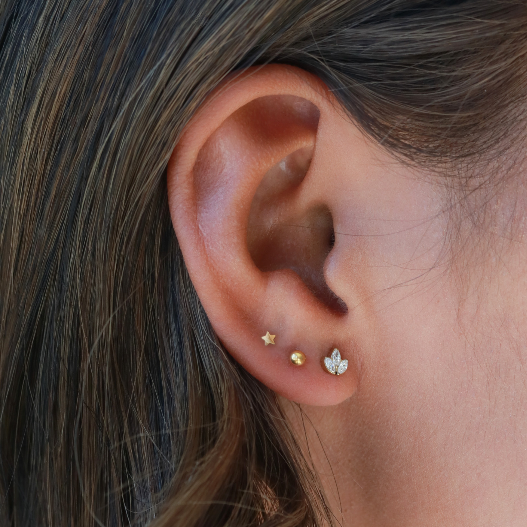 A close-up view of a person's ear adorned with three small gold earrings, each featuring a different design.
