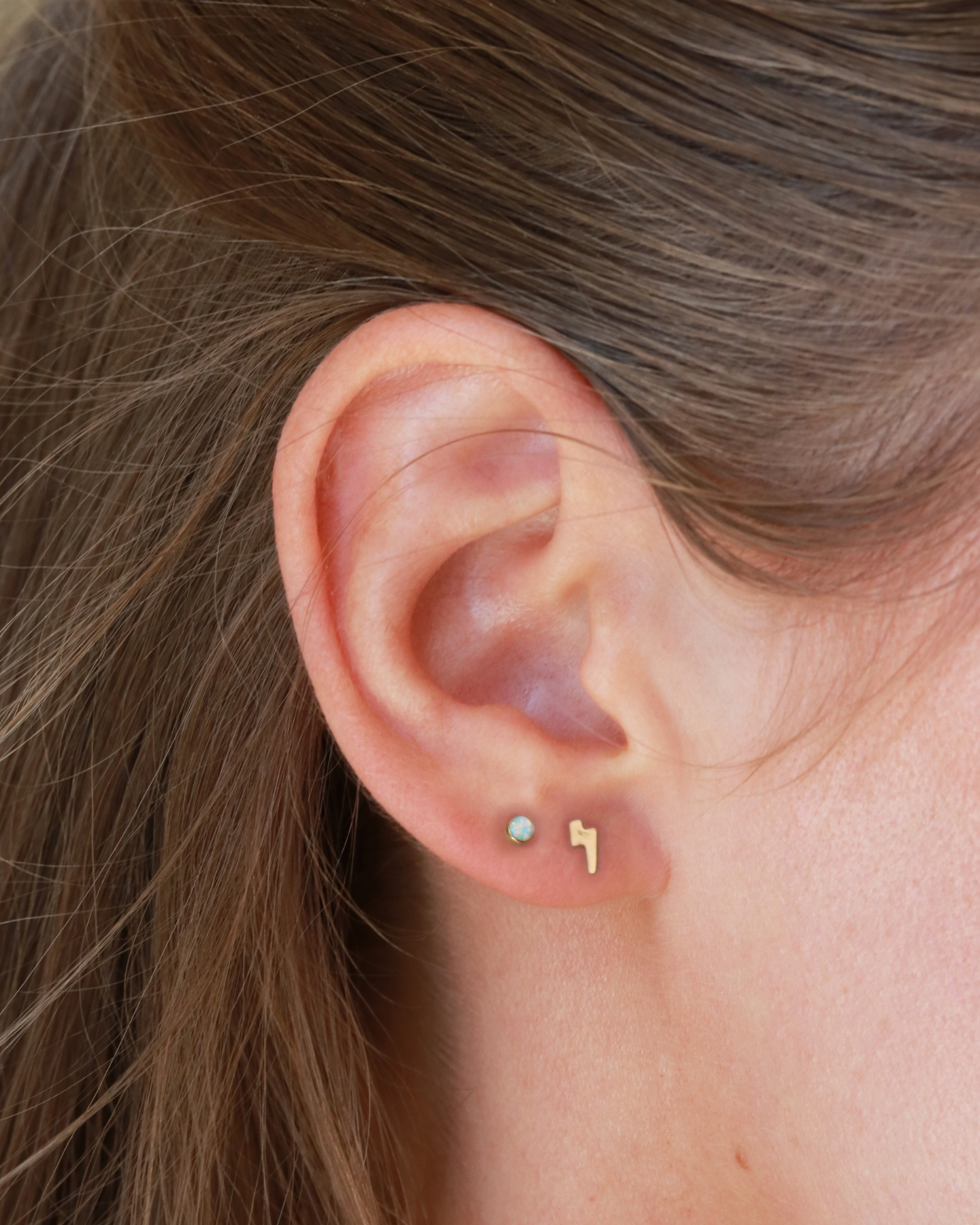 A close-up view of a person's ear with two small gold earrings, one shaped like a lightning bolt and the other a heart-shaped charm.