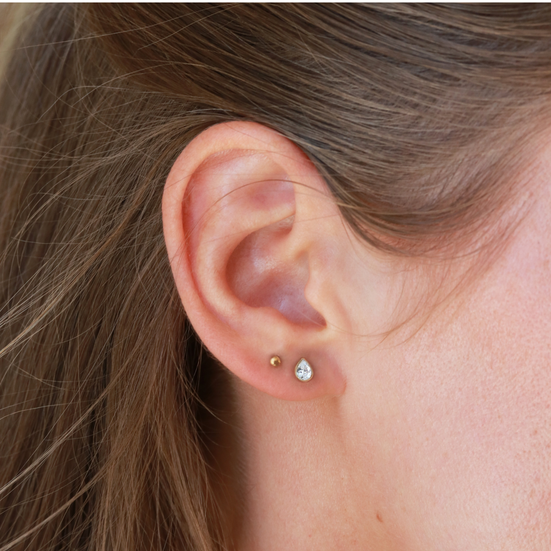 A close-up view of a person's ear with a pair of earrings, specifically a pair of earrings with a small, clear gemstone.