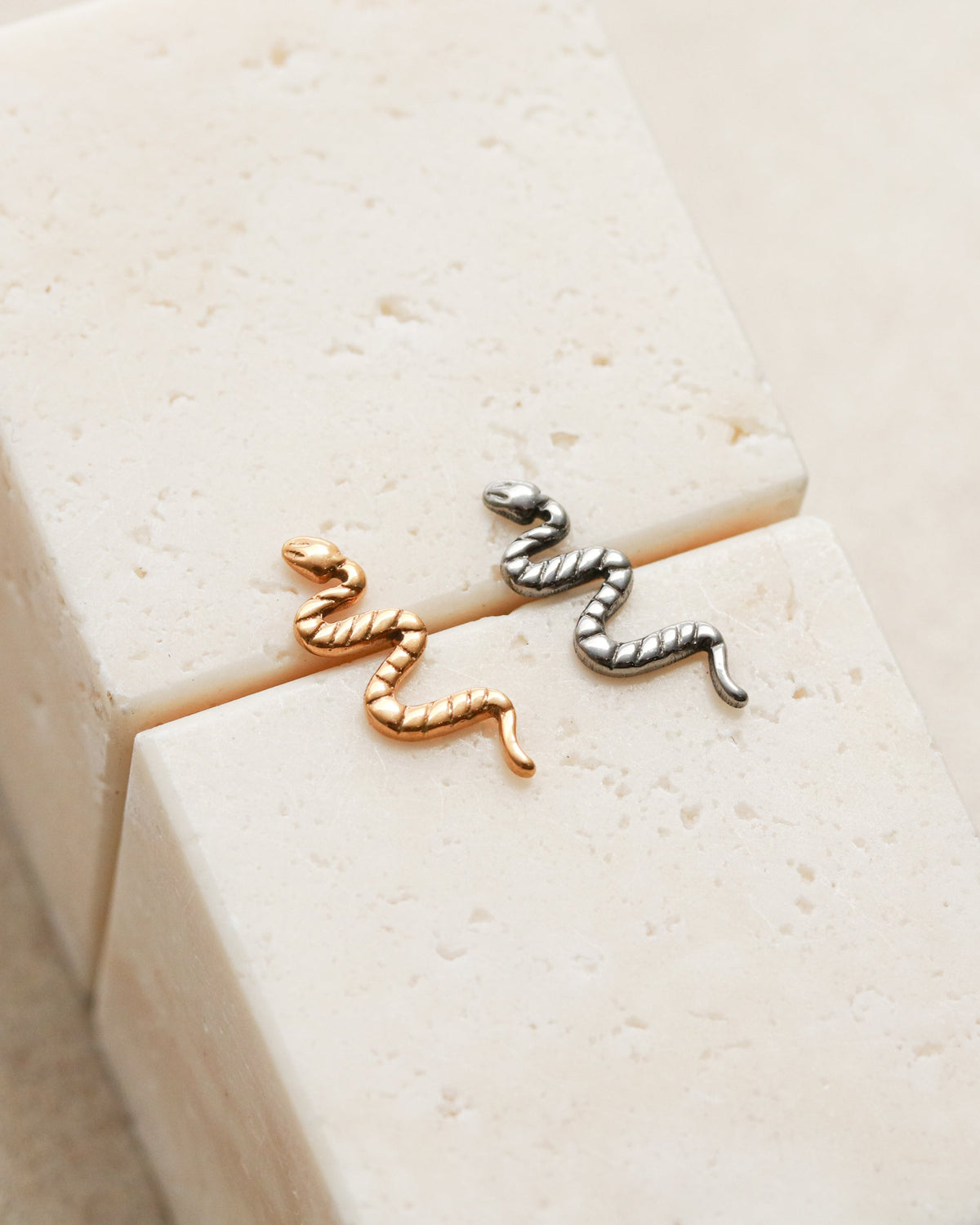 Two small metal snake-shaped earrings are placed on a white surface.