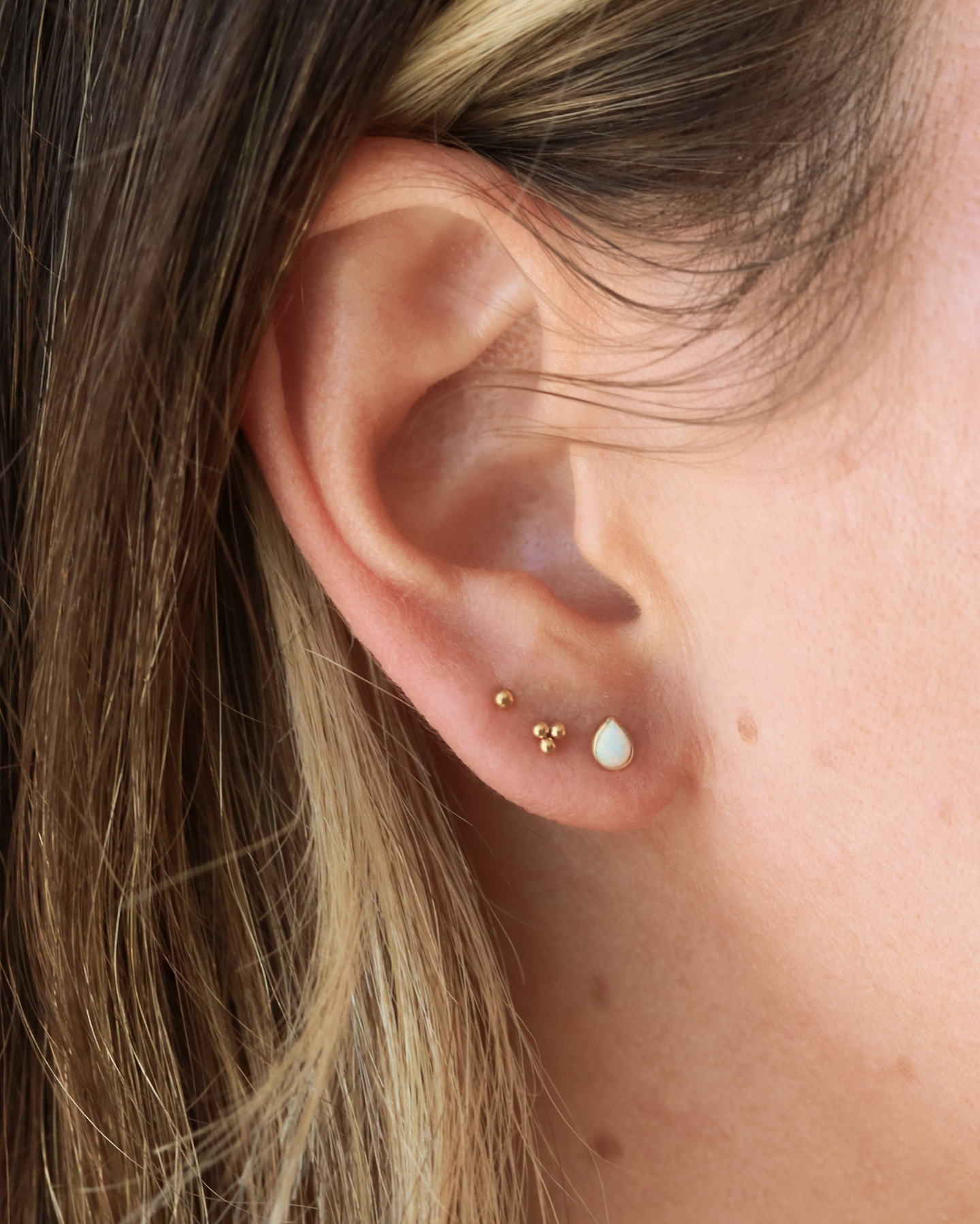 A close-up view of a person's ear adorned with two small gold earrings, one with a small white stone and the other with a small gold bead.