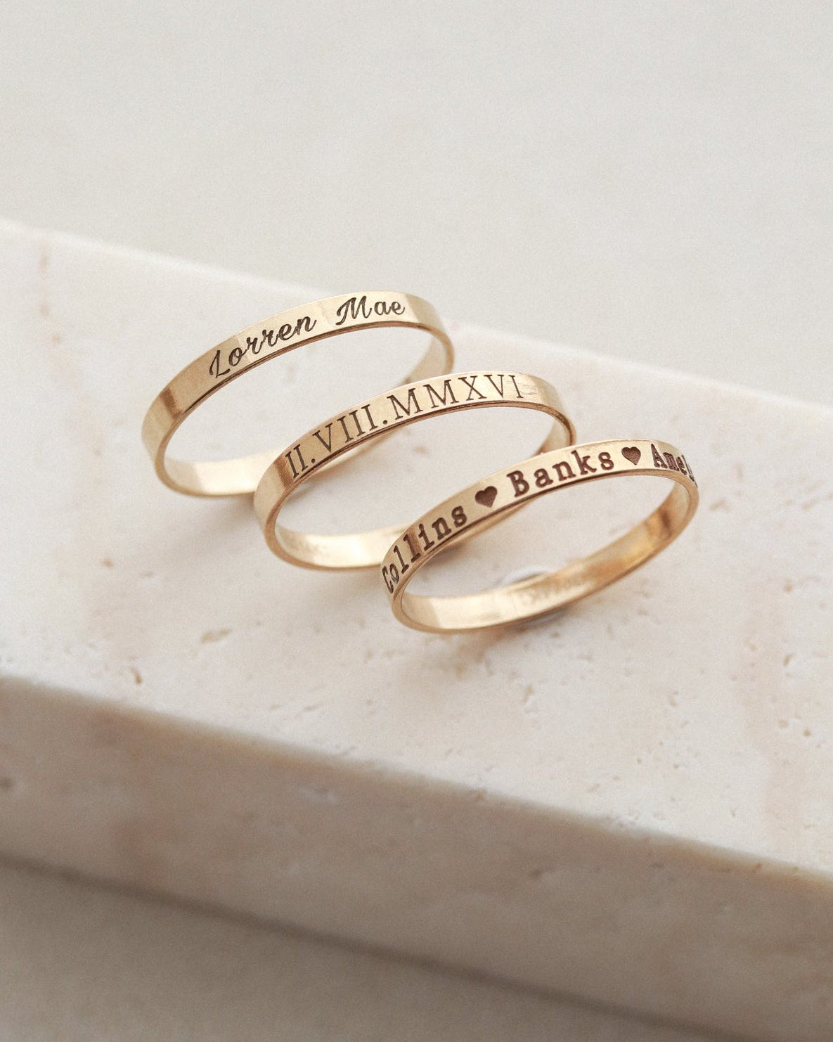 Three personalized gold rings with Roman numerals and names engraved, displayed on a light beige stone block.