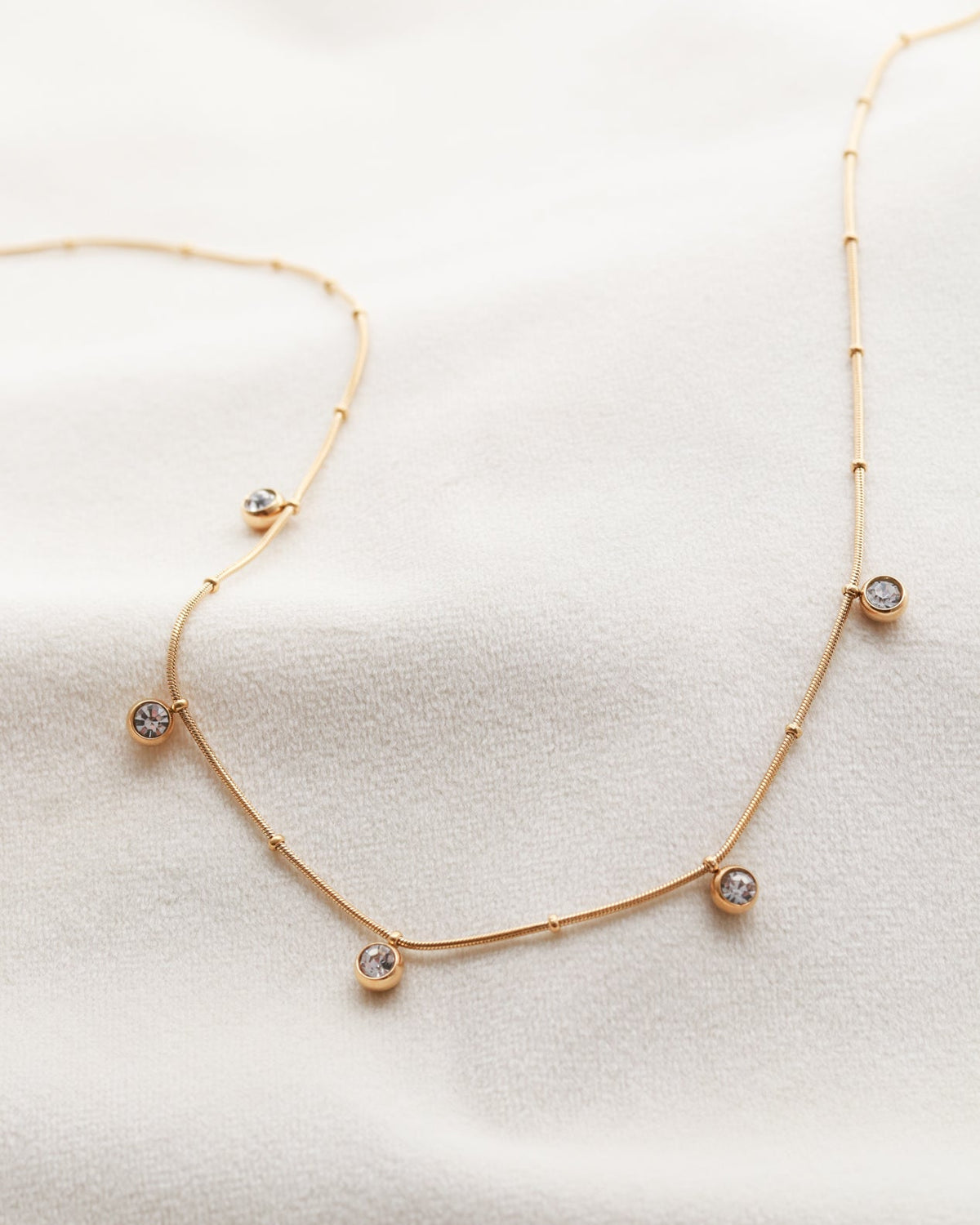 A delicate gold necklace with small, clear gemstones dangles from a white fabric background.