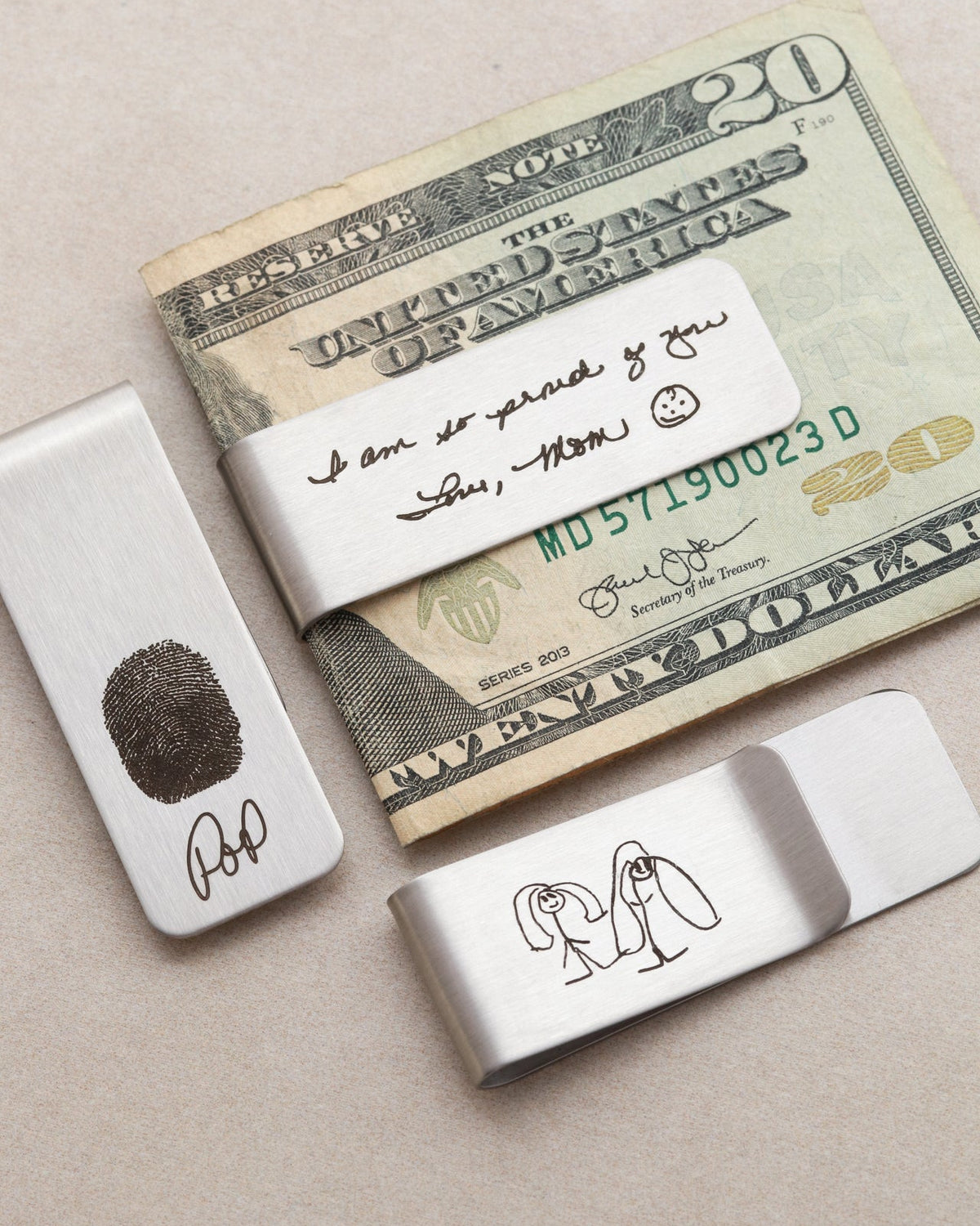 Two silver money clips with engraved designs on a piece of paper resembling a $20 bill.