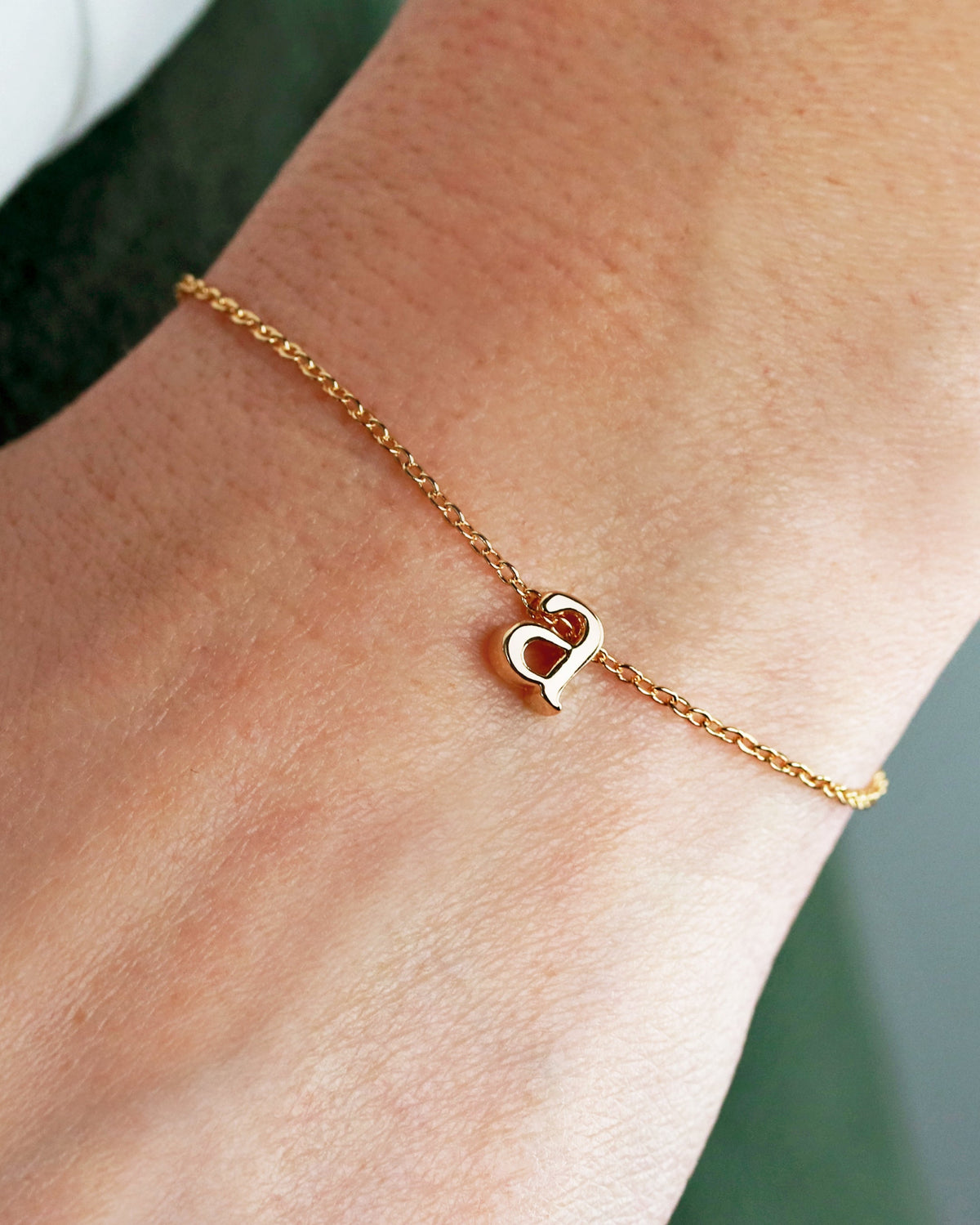 A delicate gold-plated bracelet with a small lowercase letter charm, worn on a person's wrist.