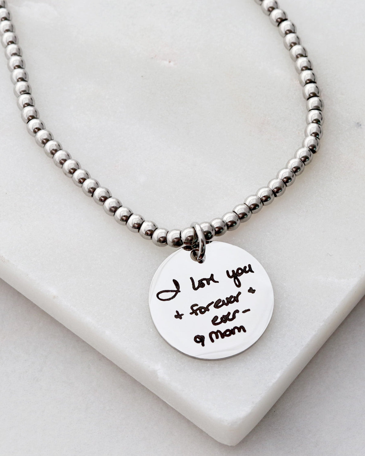 A beaded bracelet with a stainless steel chain and a personalized engraved disc pendant with handwriting.