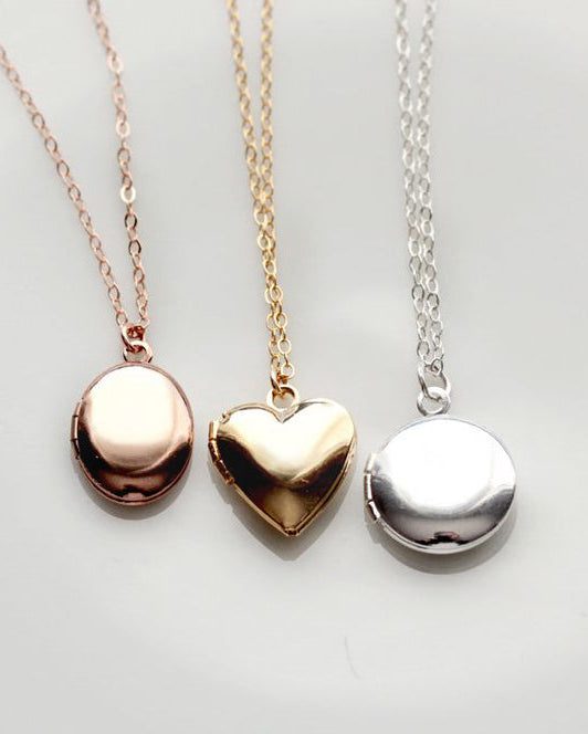 Three locket pendants on chains, in gold, silver, and rose gold colors, corresponding to heart, circle, and oval shapes, displayed against a white background.