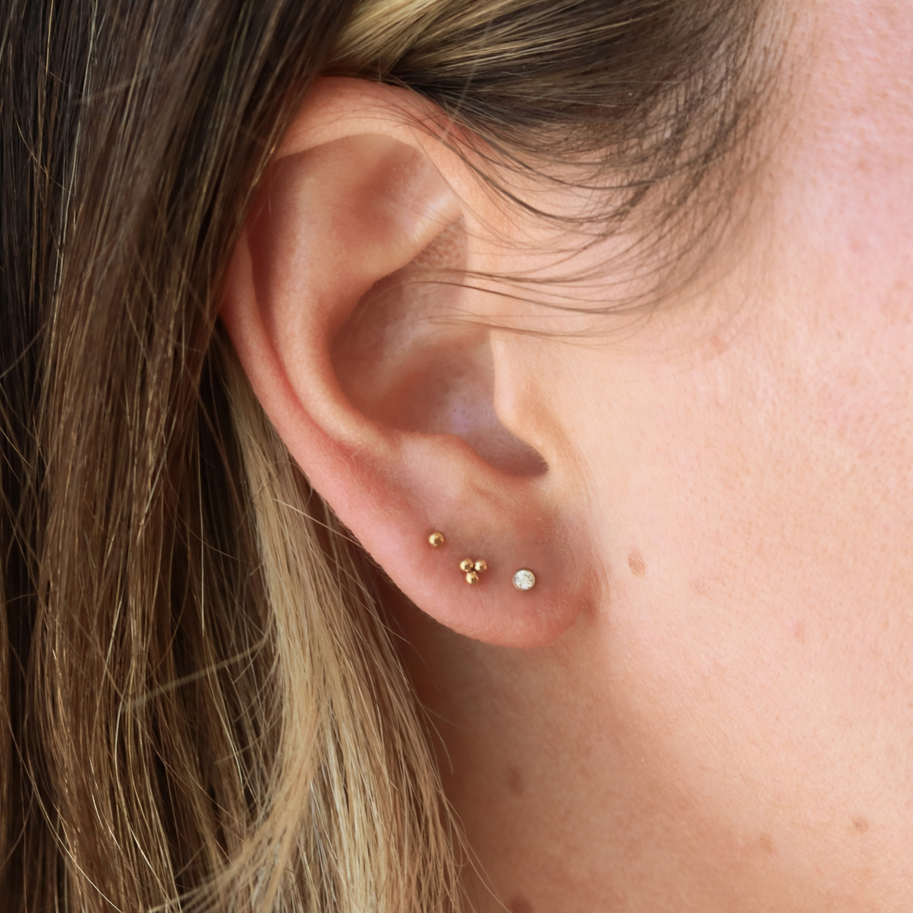 A close-up view of a person's ear with three small gold earrings on the earlobe.