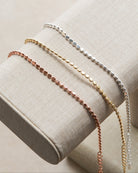 Three delicate beaded chain bracelets in silver, gold, and rose gold, displayed on a beige fabric jewelry stand.
