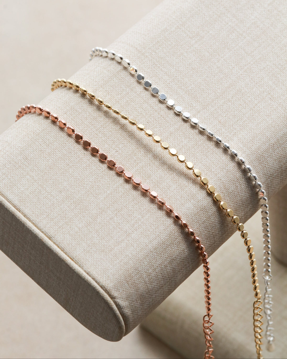 Three delicate beaded chain bracelets in silver, gold, and rose gold, displayed on a beige fabric jewelry stand.