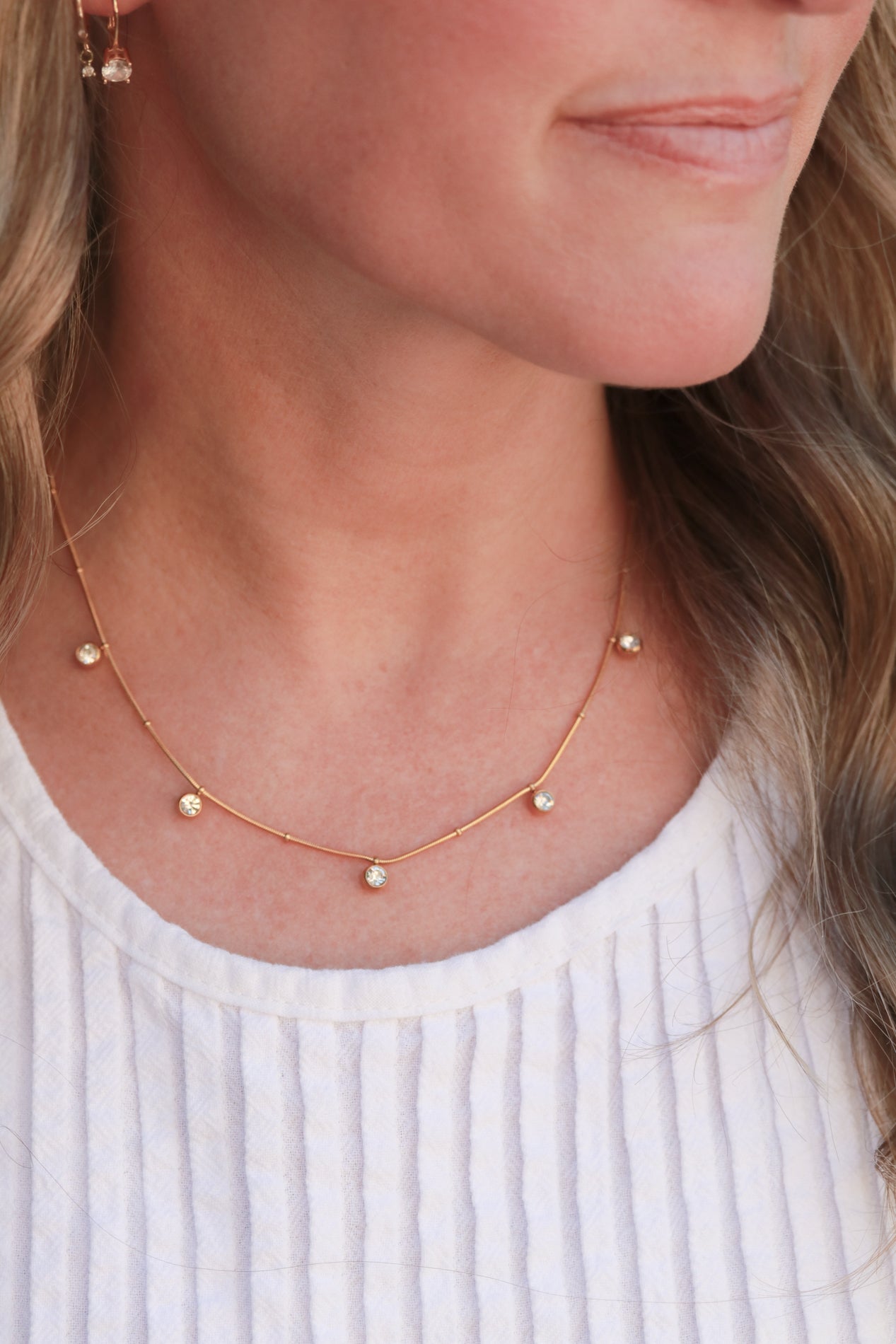 A close-up of a woman's neck and chest area, featuring a delicate gold necklace with small, clear gemstones.