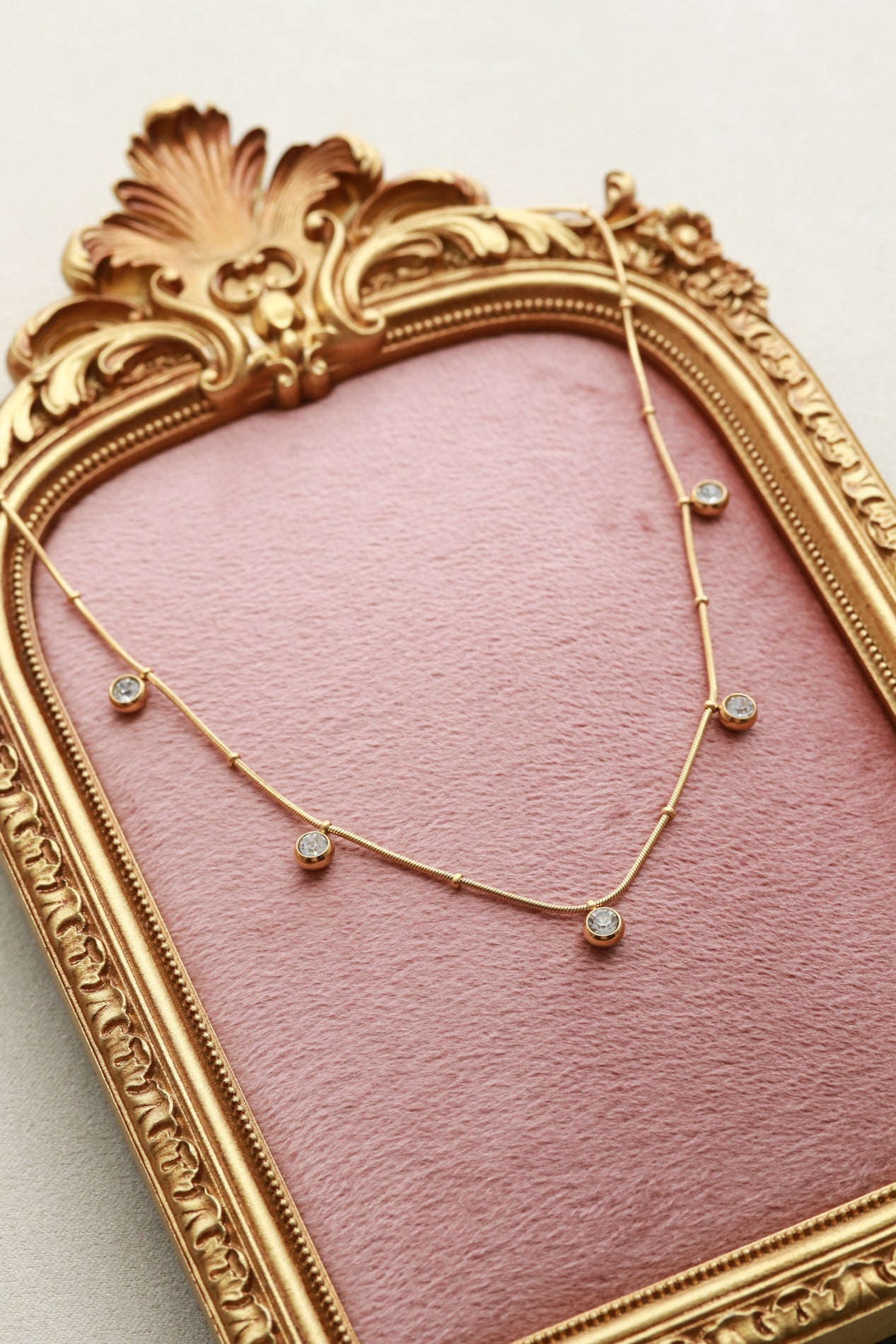 A gold-framed necklace with small diamonds is displayed on a pink velvet background.