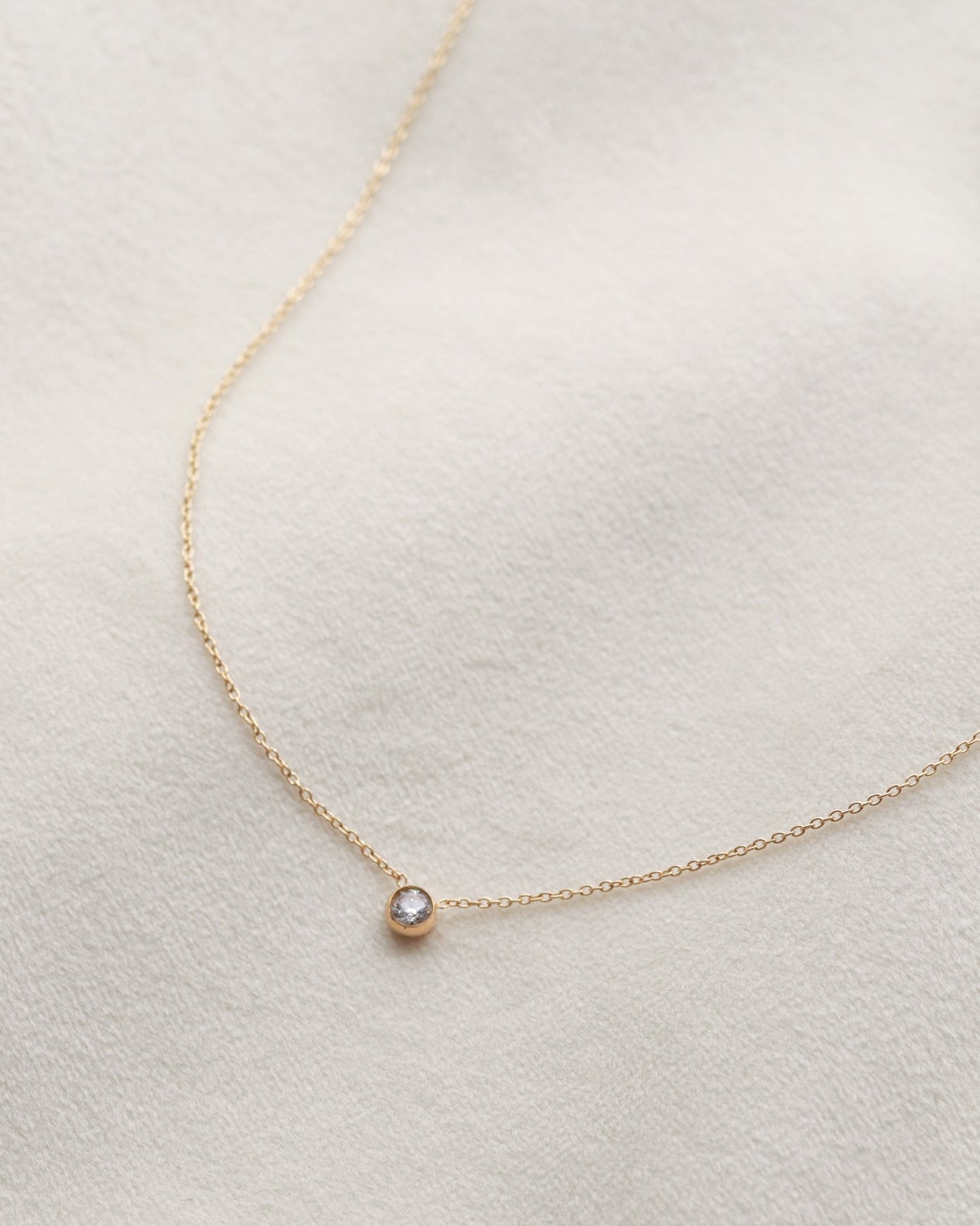 A delicate gold necklace with a small, round solitaire gemstone pendant is displayed against a white background.