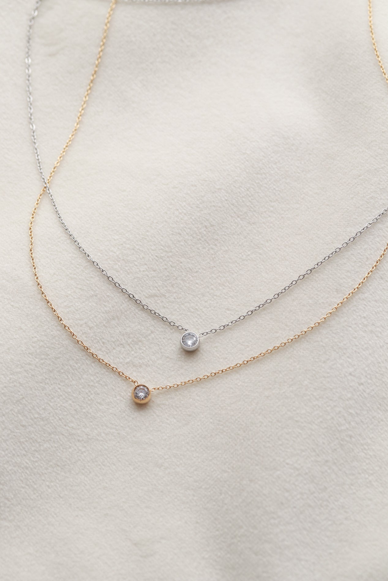 two delicate necklaces with small, clear gemstones, one in gold and one in silver, resting on a white fabric background.