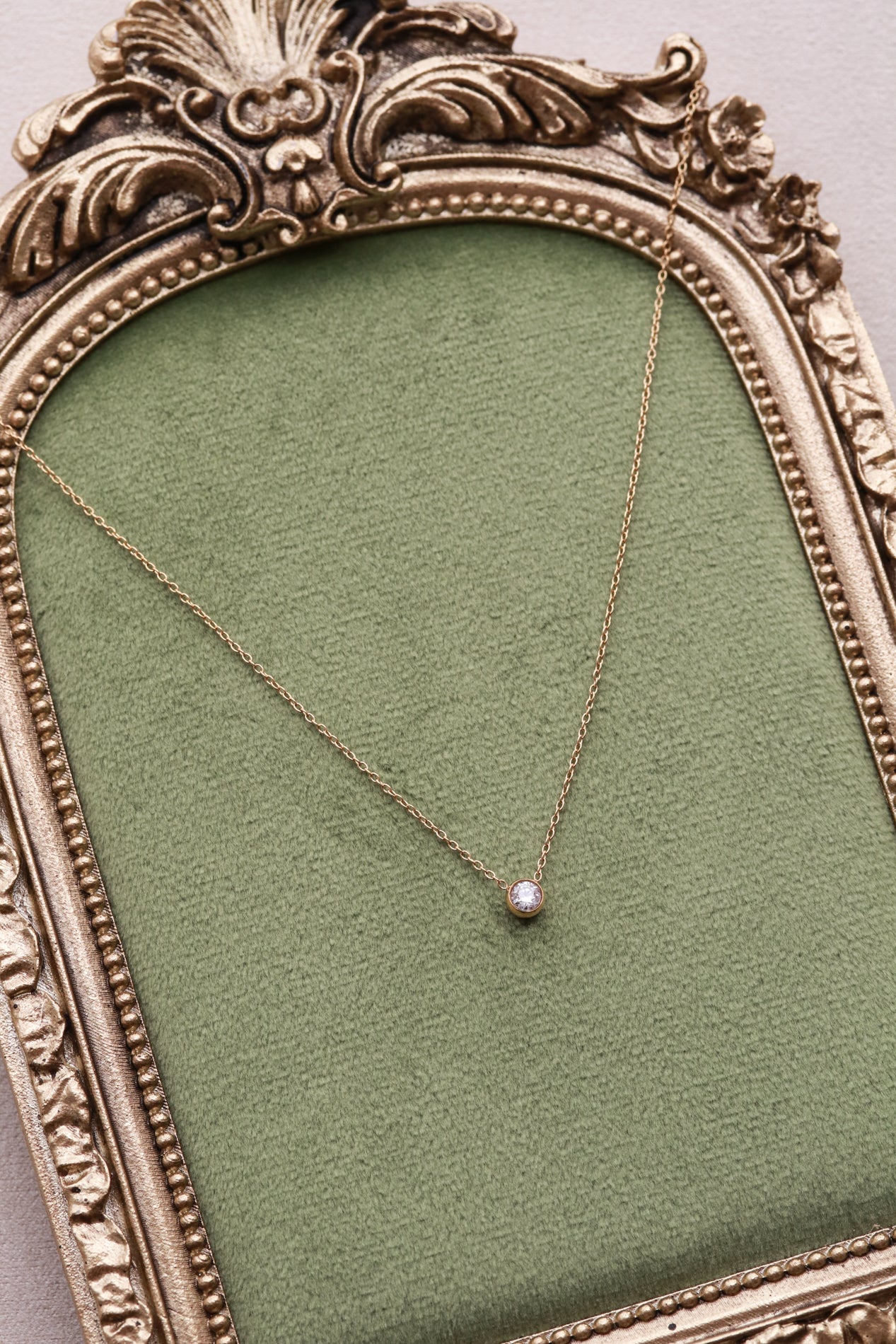 A delicate gold necklace with a small diamond pendant is displayed in an ornate gold frame against a green velvet background.