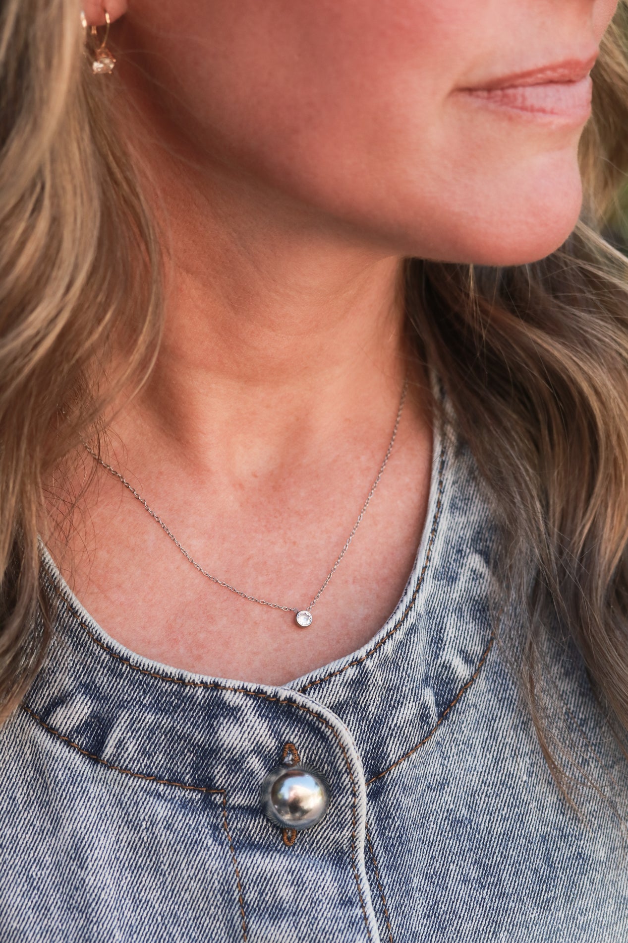 A close-up of a woman's neck and chest area, featuring a delicate necklace with a small solitaire pendant and a denim-style button on the woman's shirt.