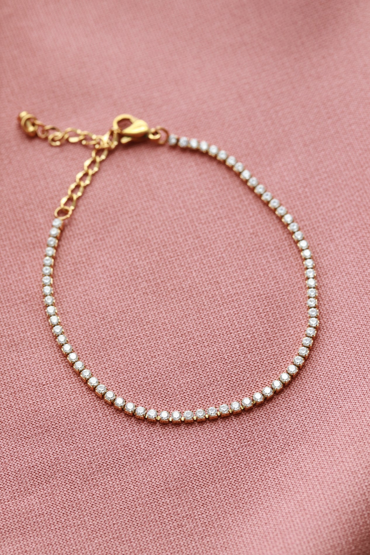 A delicate gold-colored bracelet with small diamonds adorns a pink fabric background.