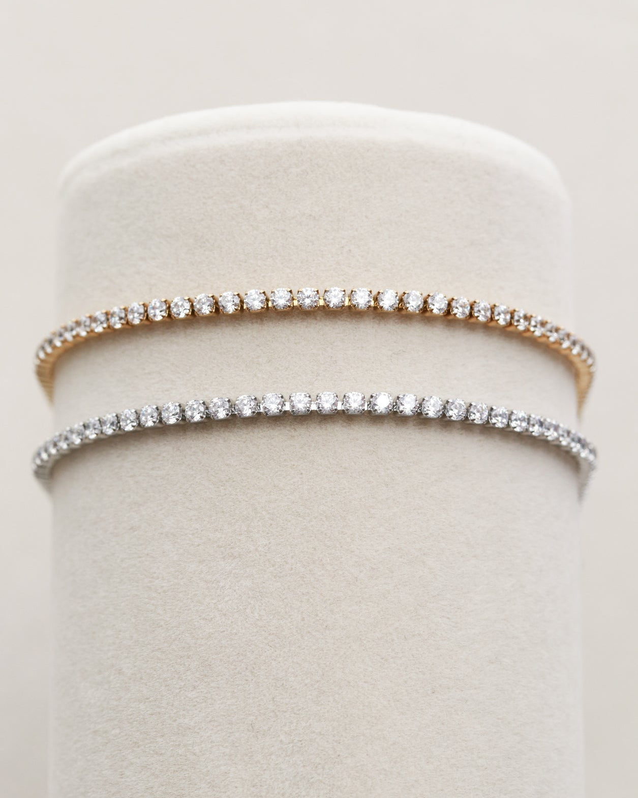 Two cz tennis bracelets with clear gemstones are displayed on a white fabric stand.