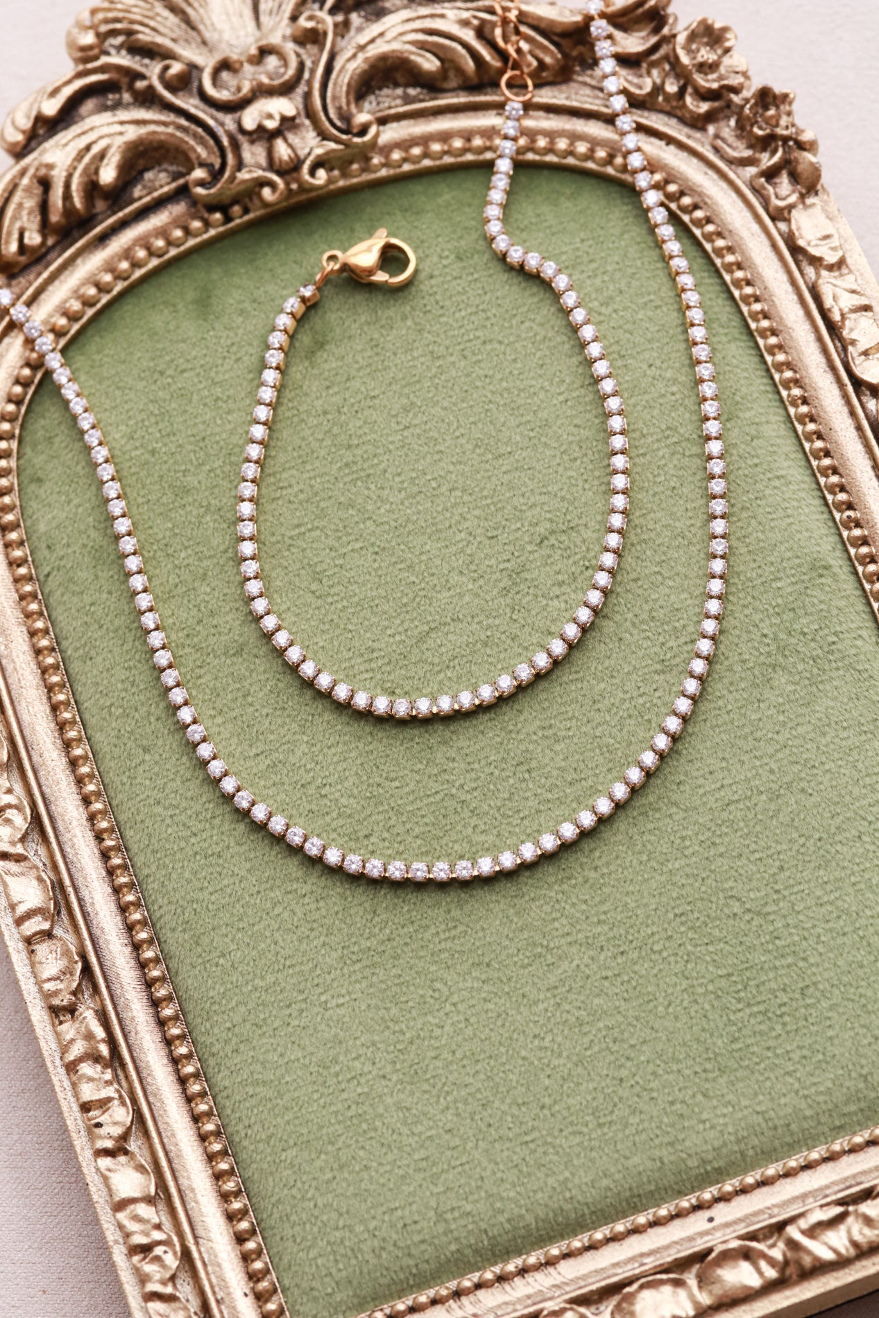 A pair of gold-colored diamond-encrusted necklaces and a bracelet are displayed on a green velvet background, placed on a decorative gold frame.