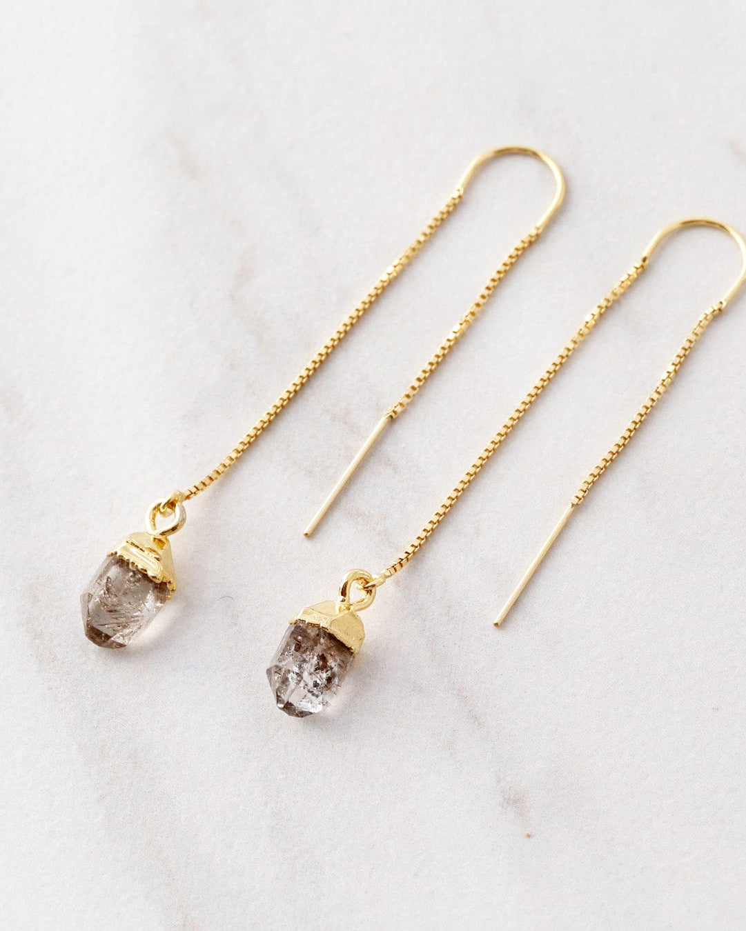 A pair of threader earrings featuring natural gemstones attached to delicate gold-colored metal chains, displayed against a marble background.