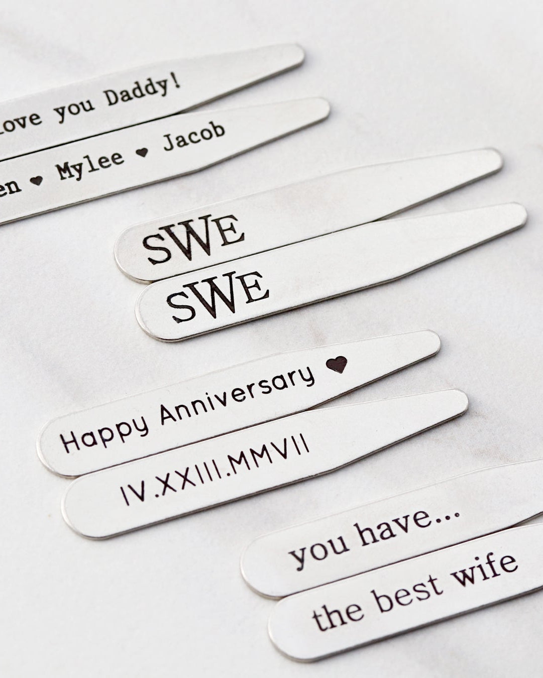 A selection of stainless steel collar stays with various engravings, placed on a marble surface.
