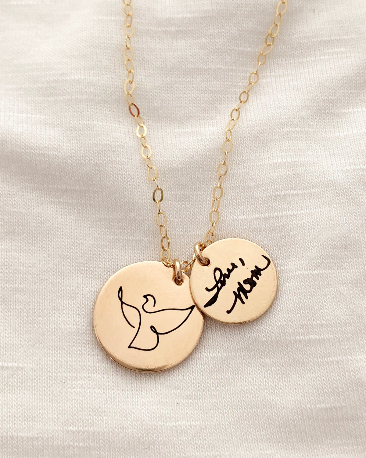 A gold necklace with two circular discs, one engraved with a dove and the other with a custom handwriting, laid on a textured fabric.