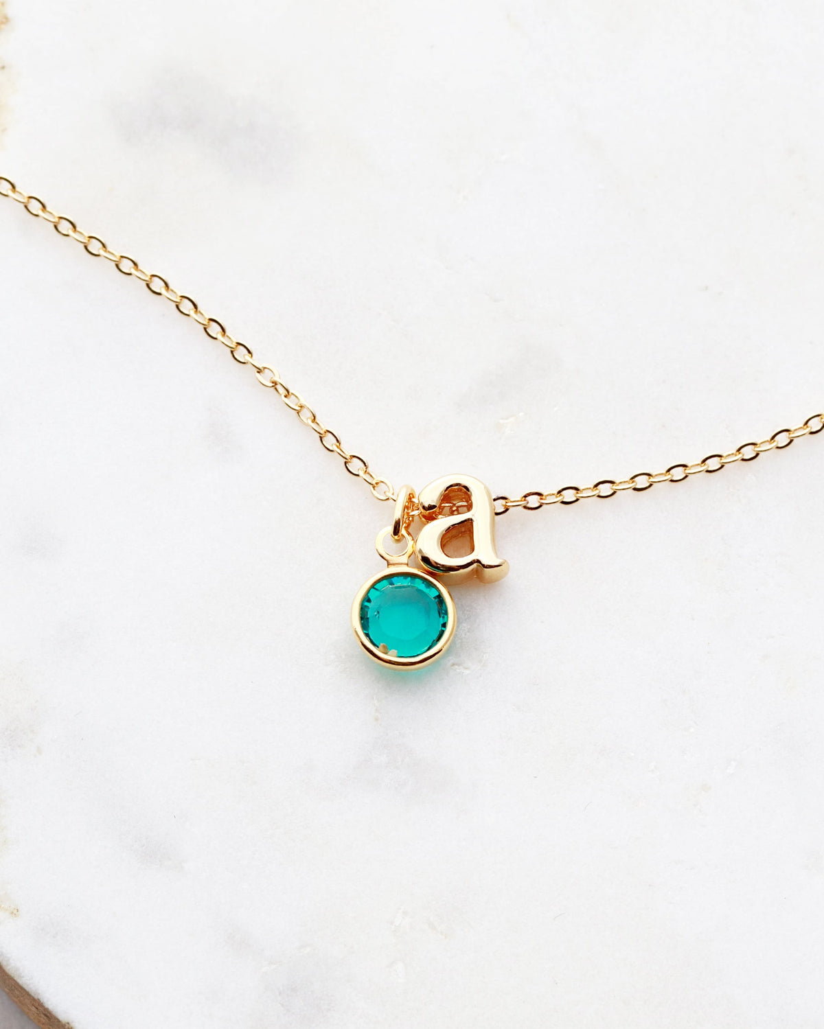 A gold-plated necklace with a round birthstone charm and a initial charm, displayed on a marble surface.