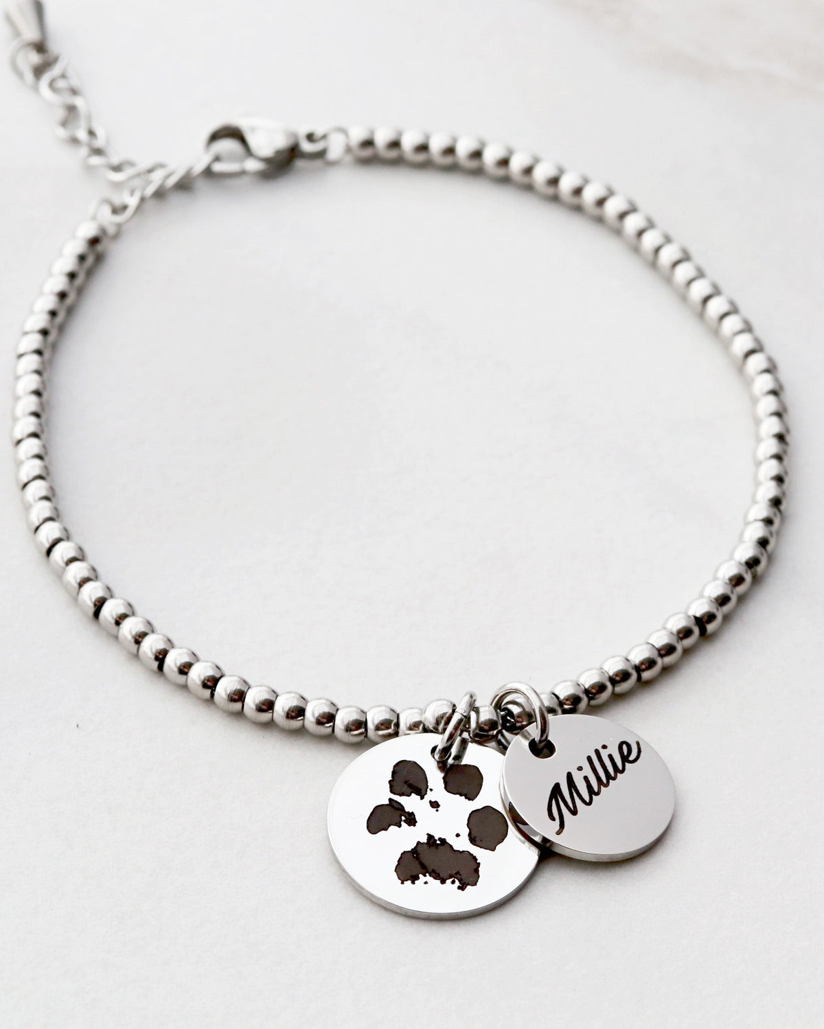 A stainless steel bracelet with beaded chain and two engraved discs, one with a paw print and the other with a name.