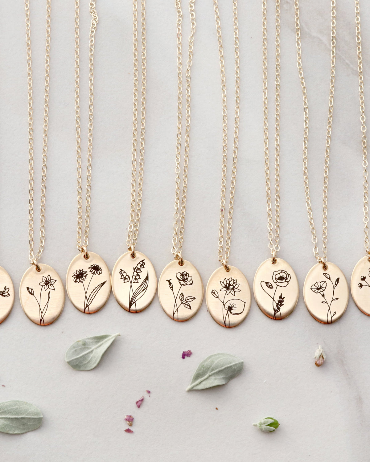A row of gold oval pendant necklaces with engraved wildflower designs, displayed on a light gray marble surface with scattered dried flower petals.