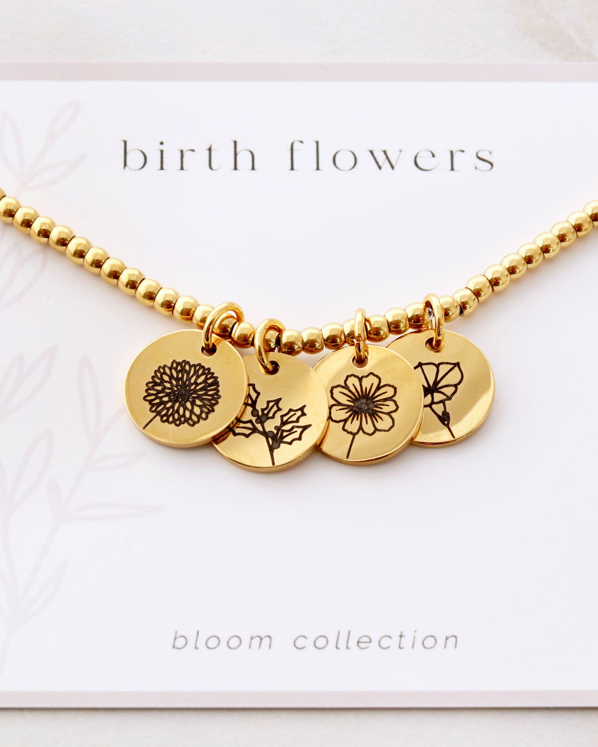 A gold-colored beaded bracelet with three engraved disc charms featuring birth flower designs, presented on a card with the text 'birth flowers' and 'bloom collection'.