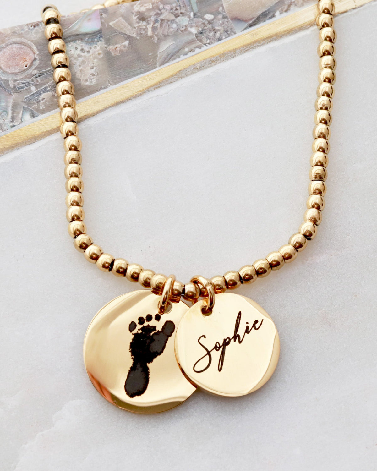 A gold-colored beaded bracelet with a custom print engraving on a pendant, displayed on a flat surface.